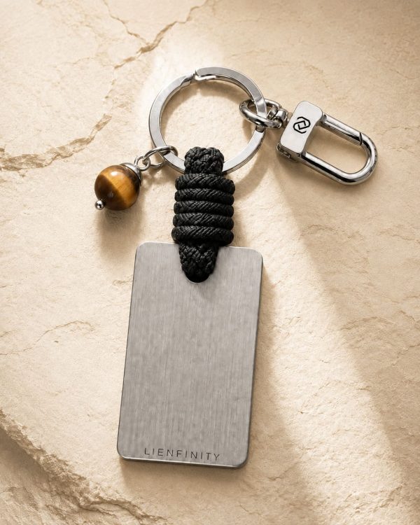 Porte-clé LIENFINITY avec œil de tigre, corde noire et plaque gravée sur fond beige texturé