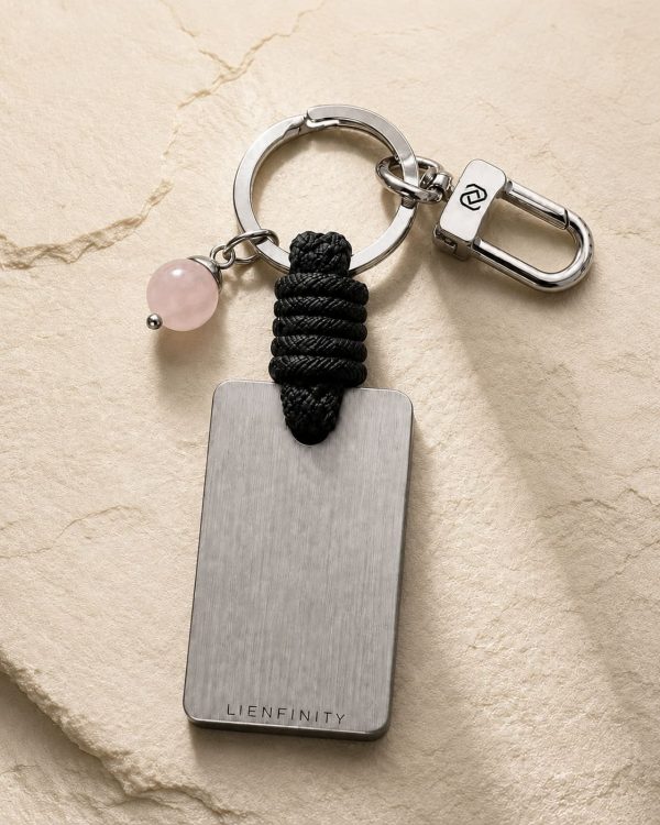 Porte-clé LIENFINITY avec quartz rose, corde noire et plaque gravée sur fond beige texturé