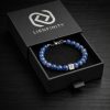 Bracelet LIENFINITY. en pierre naturelle de Lapis-Lazuli bleu intense parsemé d'éclats dorés, avec son insert de connexion en acier inoxydable 316L. Le bijou est présenté dans son écrin de luxe noir à tiroir avec le logo de la marque.