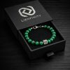 Bracelet LIENFINITY. en pierre naturelle de Malachite verte avec son insert en acier inoxydable, présenté devant son écrin de luxe noir et blanc. Le bijou met en avant les motifs circulaires naturels de la pierre et la Signature QR code gravée.