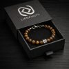 bracelet-oeil-de-tigre-boite-cadeau-lienfinity Bracelet LIENFINITY en perles Œil de Tigre marron et d'une perle en acier inoxydable gravée du logo, présenté dans sa boîte cadeau noire ouverte avec lettrage argenté.