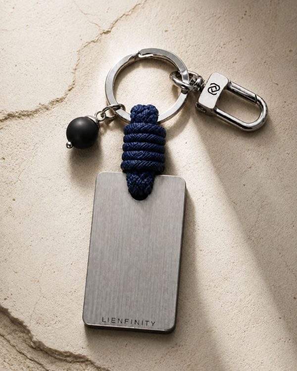 Porte-clé LIENFINITY – Onyx noir mat – Corde bleue Porte-clé LIENFINITY avec onyx noir mat, corde bleue et plaque gravée sur fond beige texturé