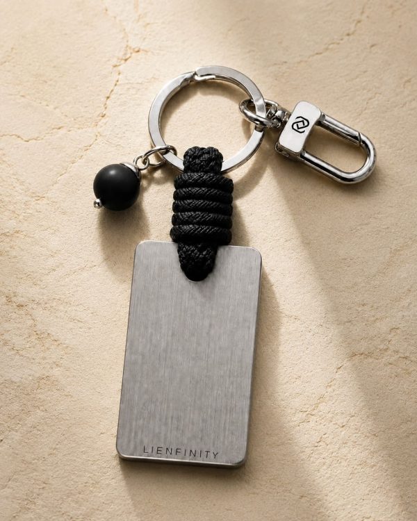 Porte-clé LIENFINITY – Onyx noir mat – Corde noire Porte-clé LIENFINITY avec onyx noir mat, corde noire et plaque gravée sur fond beige texturé