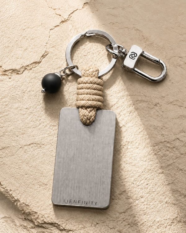 Porte-clé LIENFINITY – Onyx noir mat – Corde sable Porte-clé LIENFINITY avec onyx noir mat, corde sable et plaque gravée sur fond beige texturé